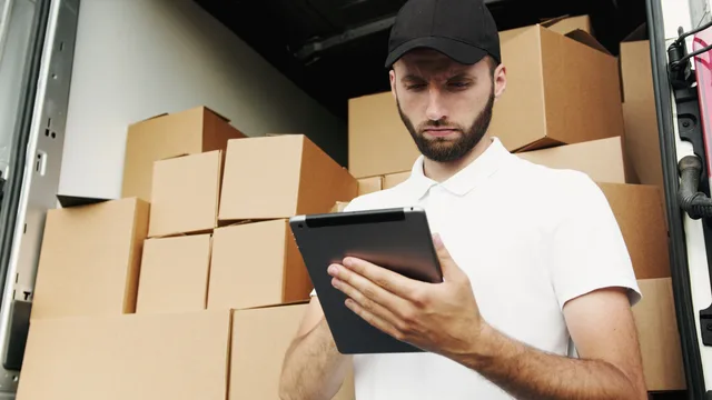Delivery worker using a tablet to manage shipments with stacked boxes in the background. Integracja ERP z systemem magazynowym: jakie dane wymieniać