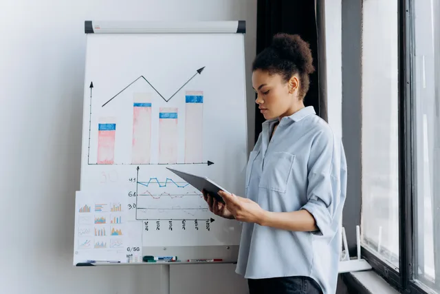 A woman analyzing sales data on a tablet next to a presentation board with graphs. Jak opisać lejek sprzedażowy przed wdrożeniem CRM